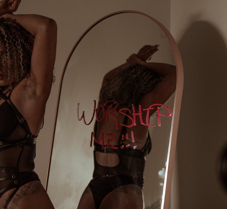 women in black lingerie hands resting on head in background worship me!! is written on mirror in red
