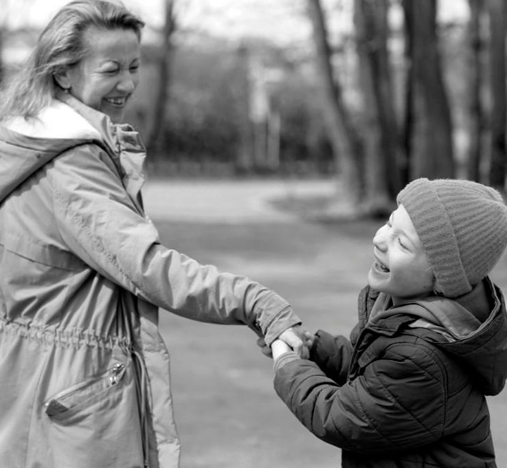 Usmiechnięty chłopiec z wadą wzroku wraz z mamą na ścieżce wśród drzew.