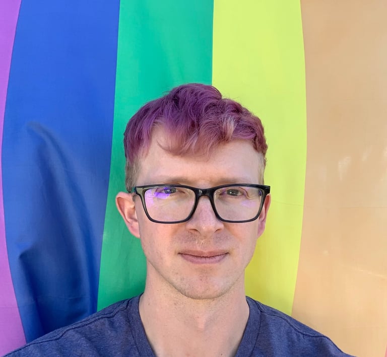 Portrait of Tomkins with purple hair with a rainbow flag as a backdrop.