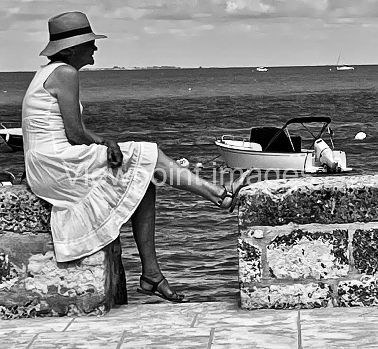 Black and white photo of a woman in a sun hat and white dress sitting on a stone wall by the sea.