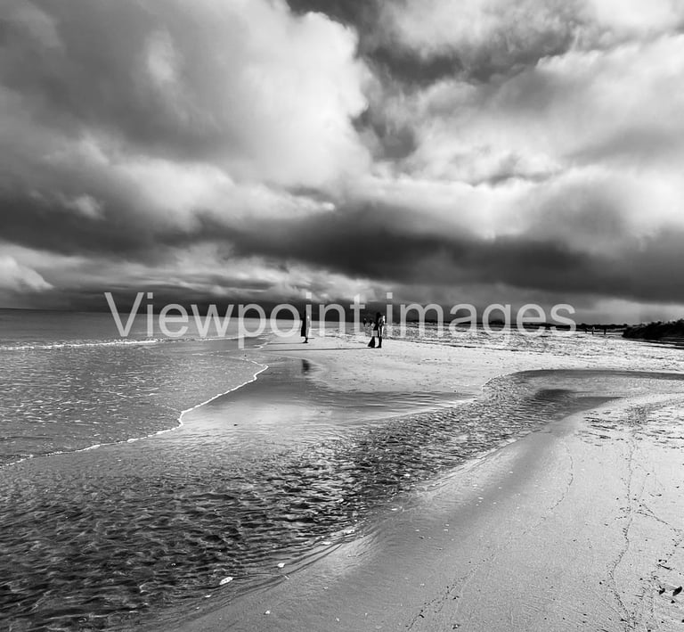 Dramatic black and white landscape of a sandy beach with dark storm clouds reflecting on the ocean tide.