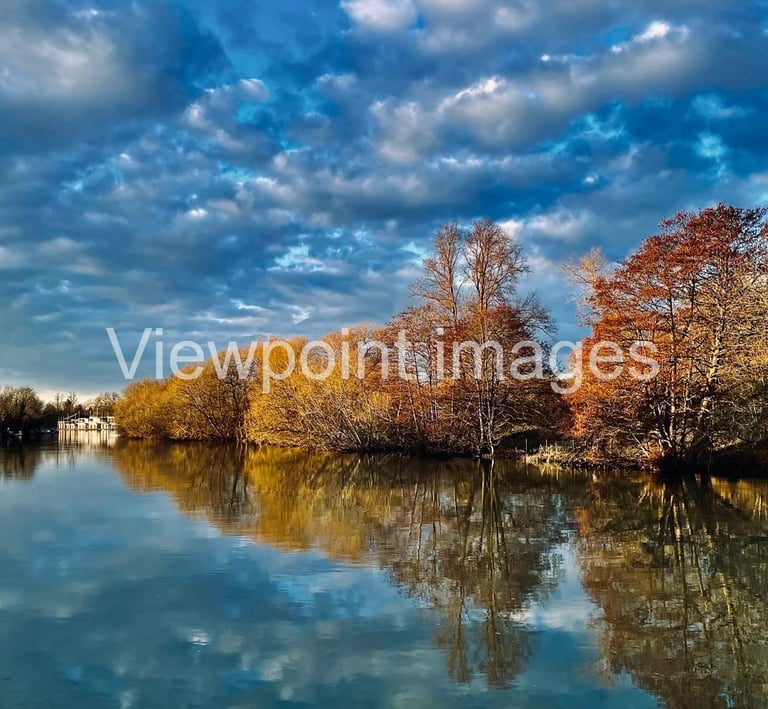 Tranquil river landscape with autumn trees reflecting in calm water under a dramatic blue sky.