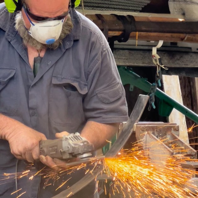 A metalworker wearing safety gear uses an angle grinder to cut metal, creating bright orange sparks.