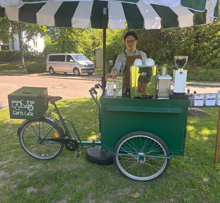 Grünes Kaffeefahrrad mit grün-weißem Schirm und Barista auf einer Wiese.