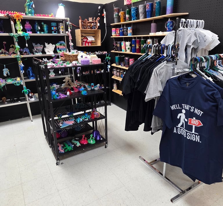 Gift shop interior with 3D printed dragons, colorful tumblers, and custom graphic t-shirts on display.