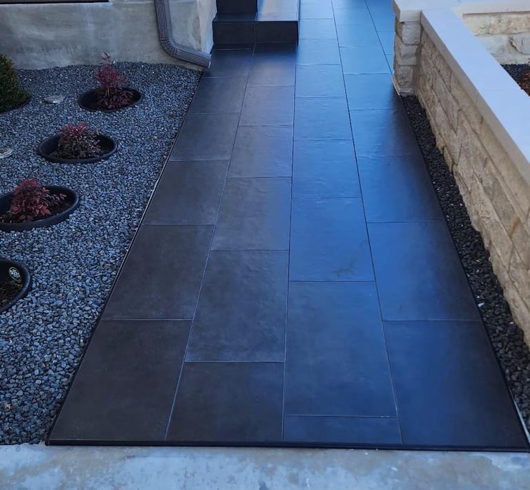 A modern dark grey porcelain tile walkway leading to a stone house entrance with xeriscape landscaping.