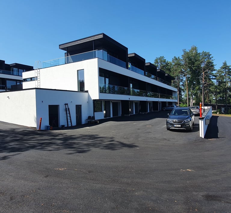 Modern white apartment building with glass balconies and paved parking area under a clear blue sky.
