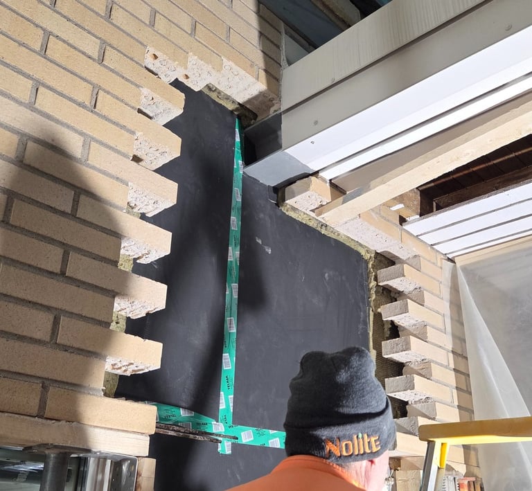 A construction worker installing black building insulation and flashing into an exterior brick wall.