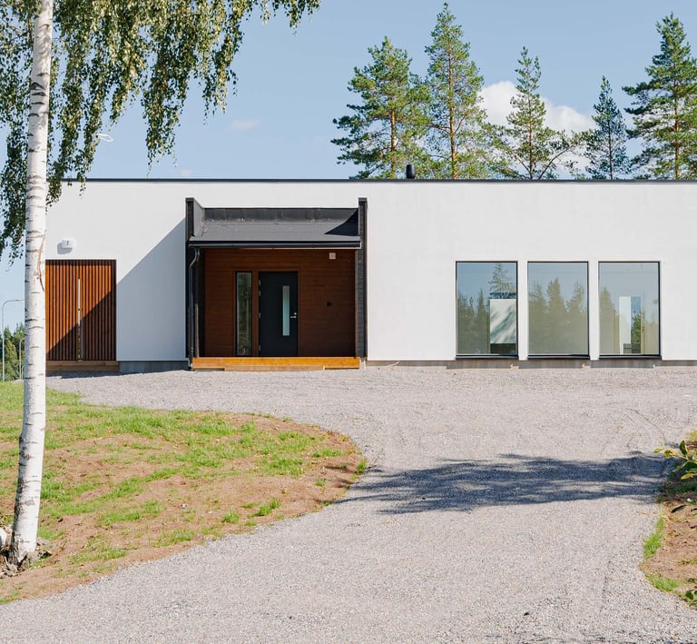 Modern white single-story house with minimalist design and gravel driveway surrounded by pine trees.