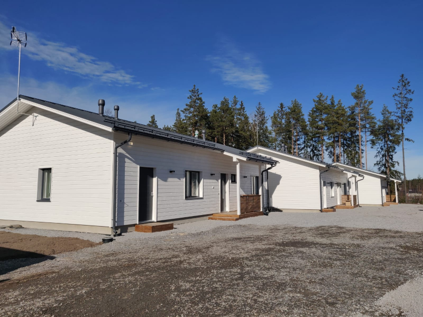 Modern white wooden row houses with gravel driveways under a clear blue sky and pine trees.