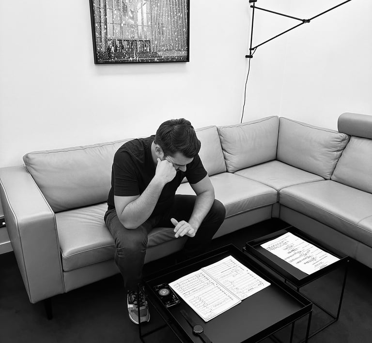 A man sits on a grey sectional sofa reviewing music sheets on a minimalist black coffee table.