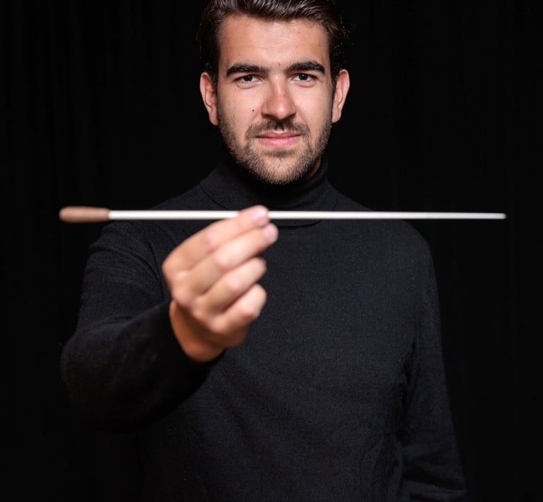 A male orchestra conductor in a black turtleneck holding a white baton against a dark background.