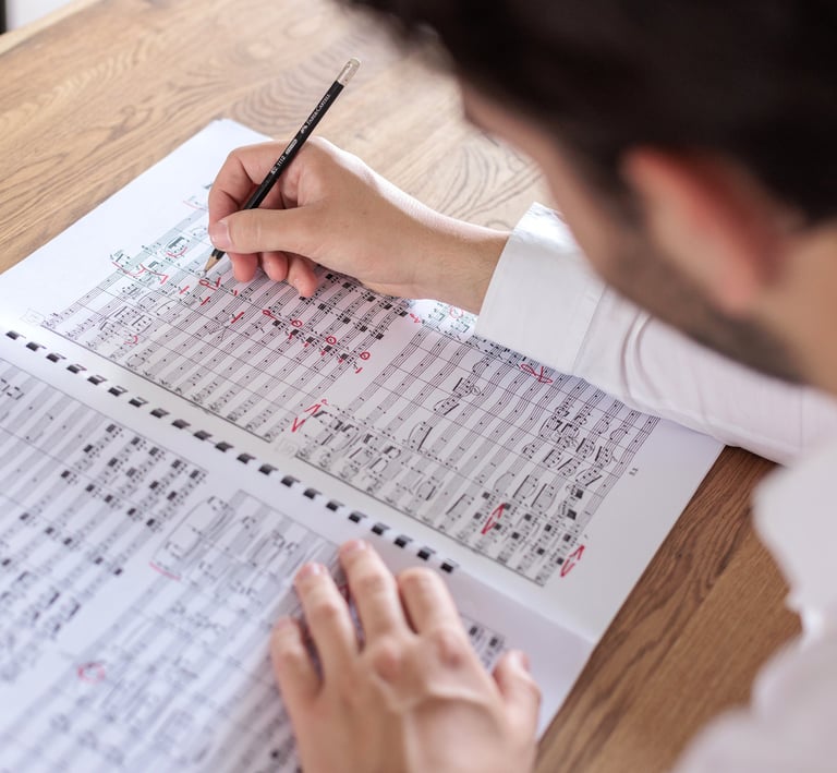 A composer writing and editing a musical score with a pencil on a wooden desk.