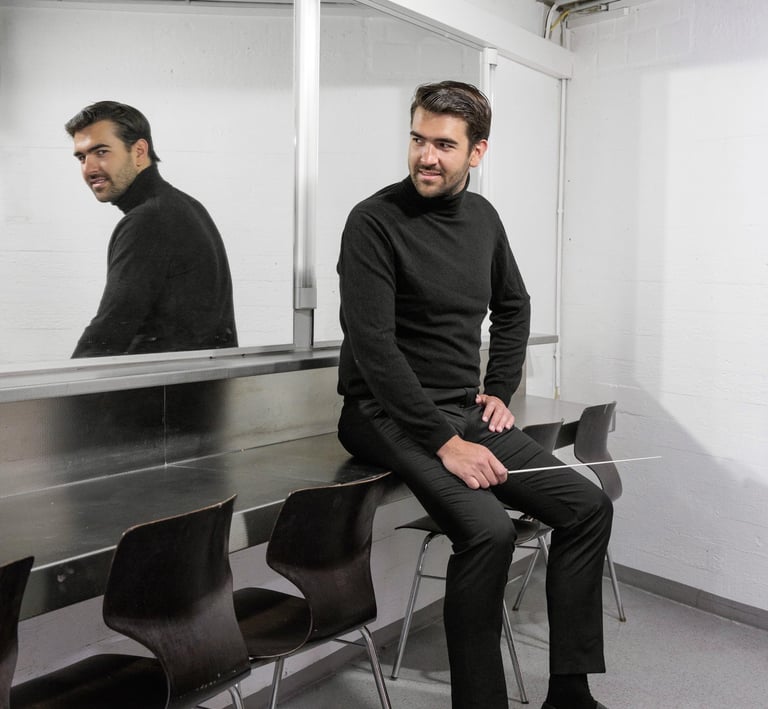 A male conductor in a black turtleneck sitting in a dressing room holding an orchestra baton.