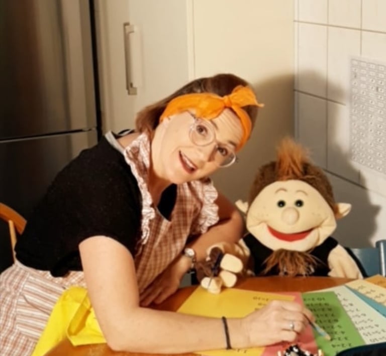 Betti Konfetti mit Handpuppe am Küchentisch erklärt die Schulaufgaben.