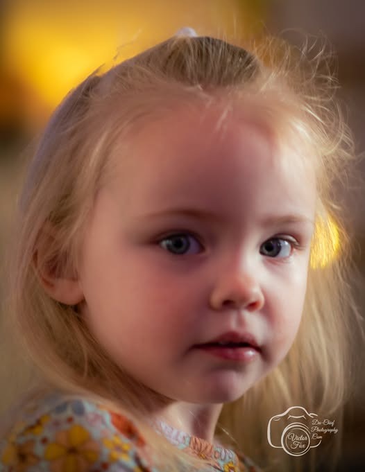 A close-up portrait of a young blonde girl with blue eyes and a floral shirt.