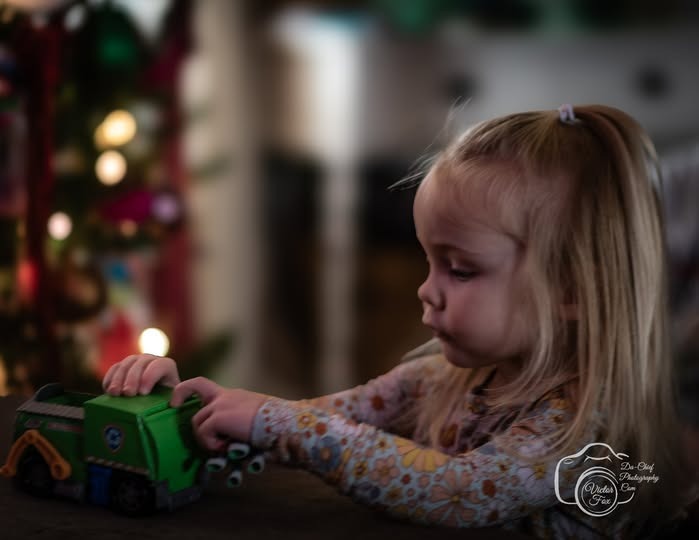 A young blonde girl playing with a green toy truck near a blurred Christmas tree.