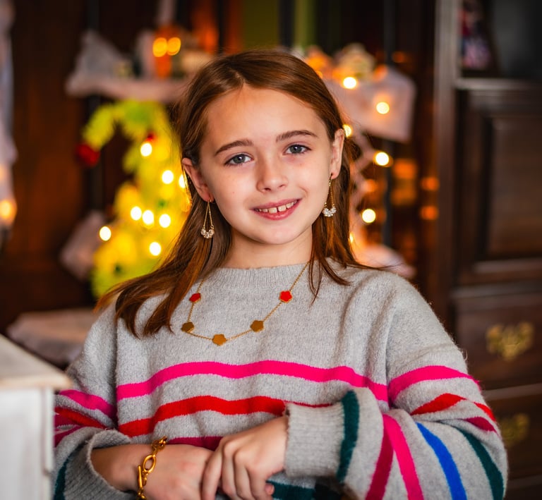 A smiling young girl in a striped sweater posing in front of festive holiday lights.