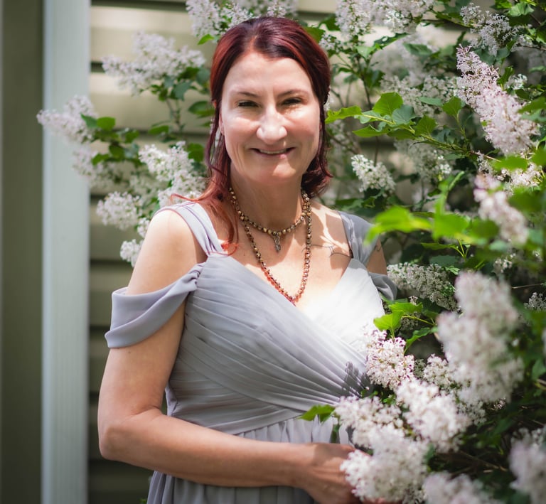 A woman in a grey formal gown smiling outdoors next to blooming white lilac flowers.