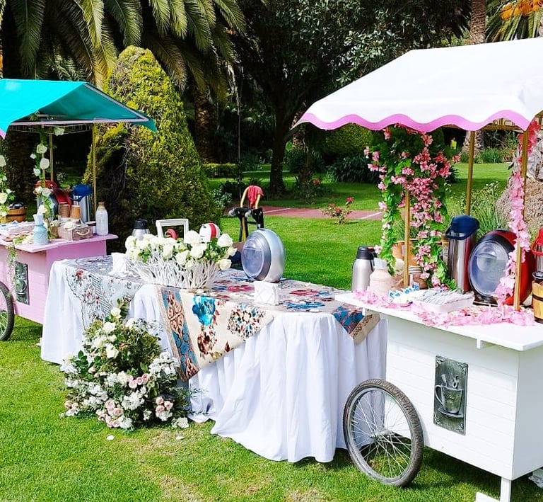 Elegant outdoor garden catering station featuring decorative white and pink beverage carts with floral arrangements.