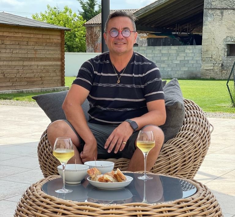 A man wearing glasses relaxes in a wicker chair outdoors with white wine and appetizers.