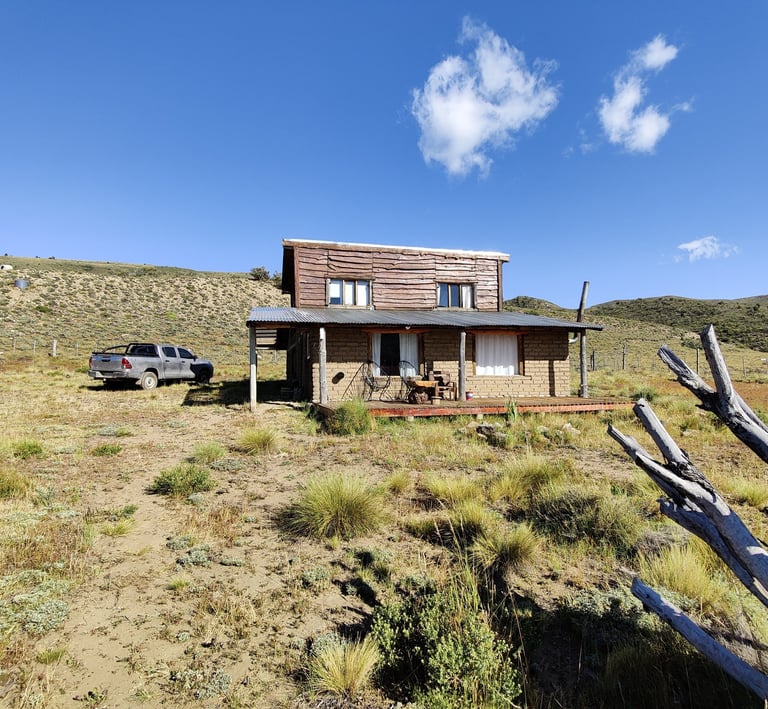 A wooden house with a metal roof on a grassy hill under a blue sky.