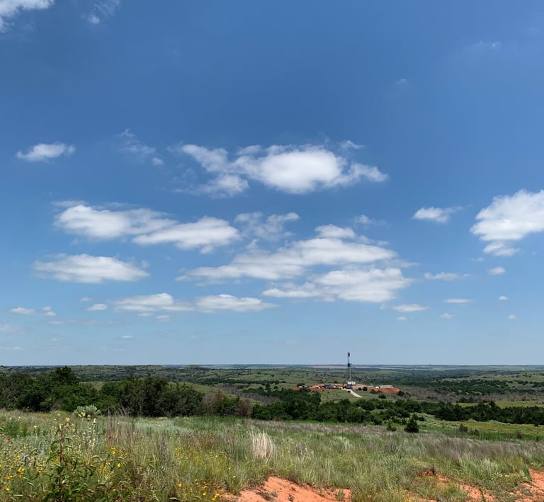 A distance view of a drilling rig on a grassy field in Oklahoma under a blue sky with clouds.