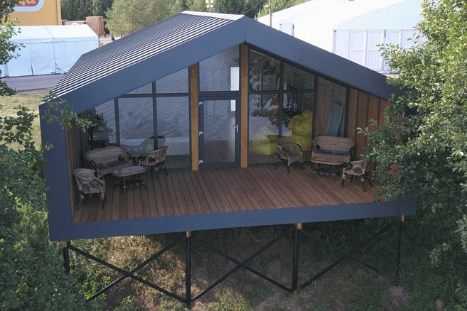 Modern black prefab cabin with glass walls and wooden deck on a metal foundation.