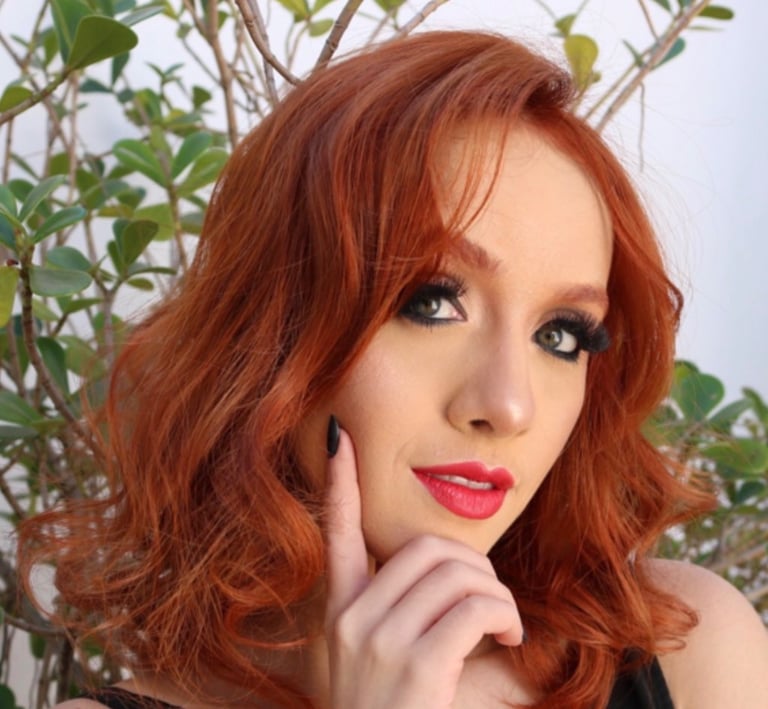 Woman with vibrant copper red hair and red lipstick posing against a green plant backdrop.