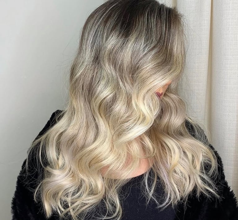 A woman with long wavy blonde balayage hair and dark roots styled in soft beach waves.