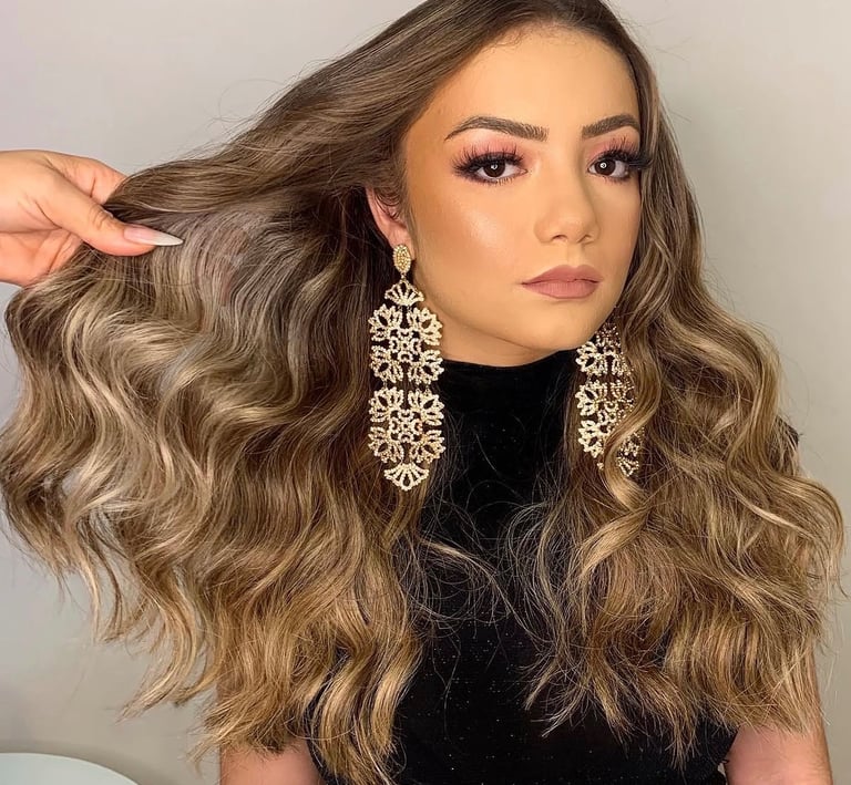 Woman with long wavy balayage hair, glamorous pink eyeshadow, and gold statement chandelier earrings.