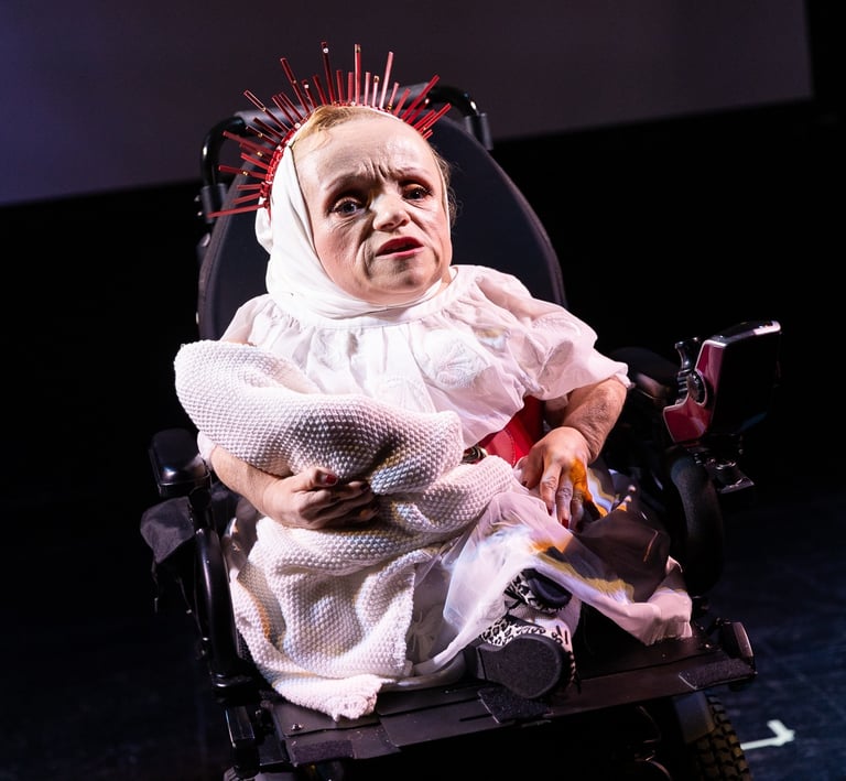 A woman of short stature wearing a white dress and crown of thorns in an electric wheelchair