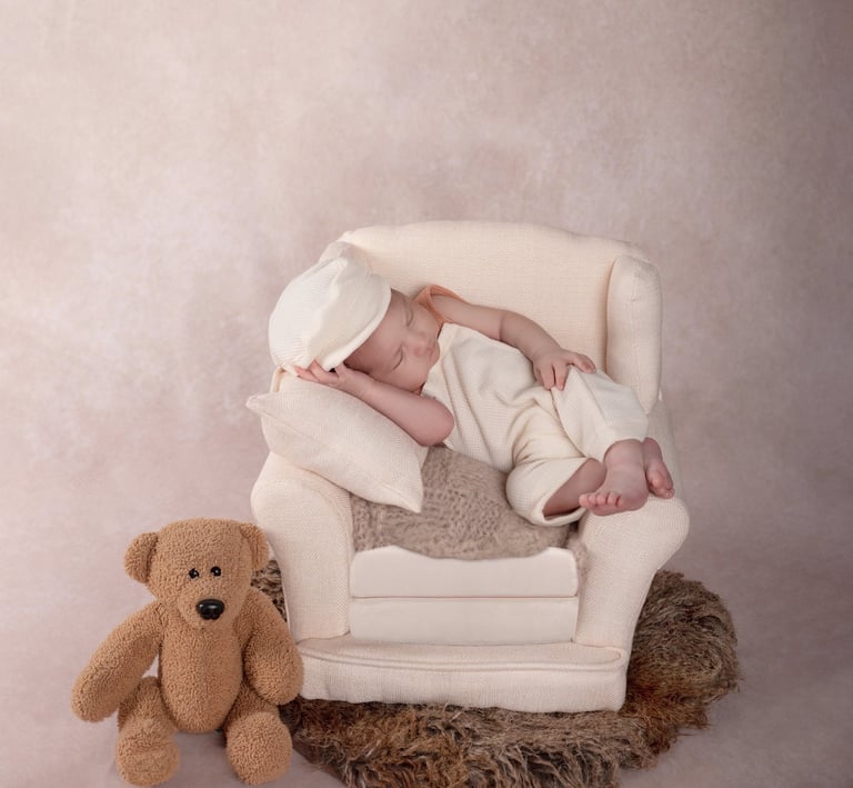 un bebé dormido en un pequeño sillón, al lado se encuentra un osito de peluche