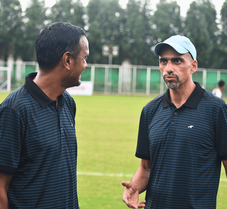 Two soccer coaches in striped polo shirts discuss strategy on a green football field.
