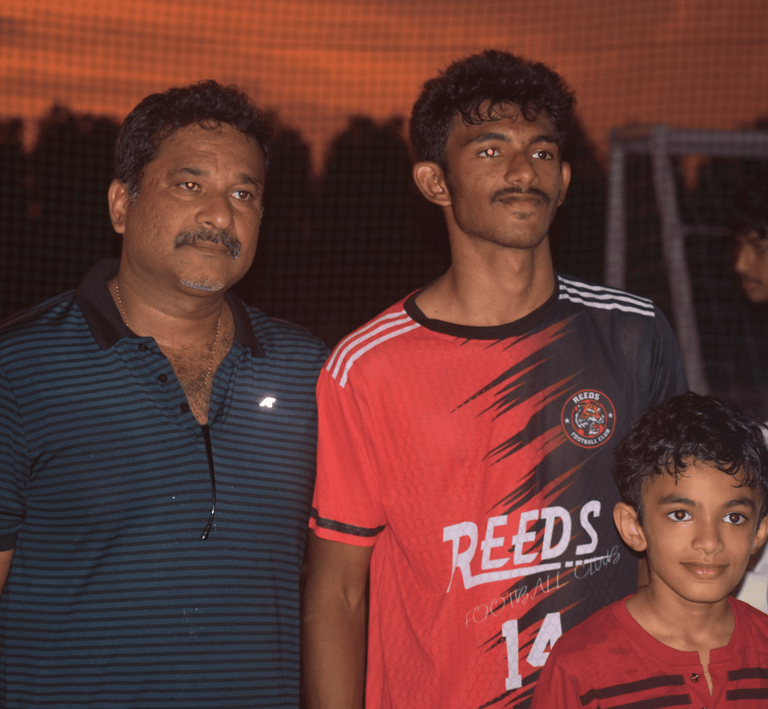 A soccer player in a Reeds Football Club jersey poses with two family members at sunset.