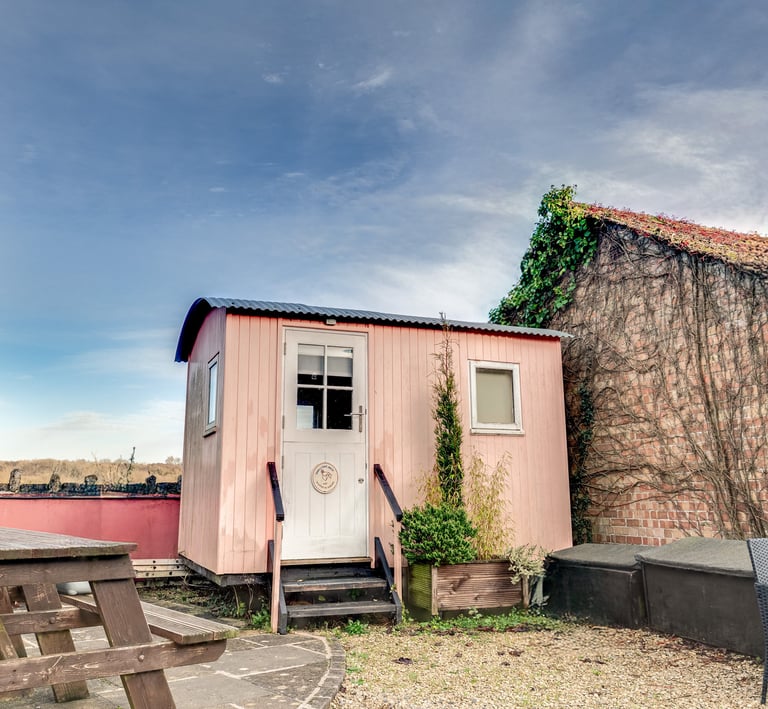 A charming pink shepherd's hut guest room with a wooden picnic table and patio seating.