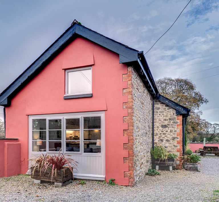 Charming red cottage with stone walls, large windows, and a gravel courtyard under a blue sky.