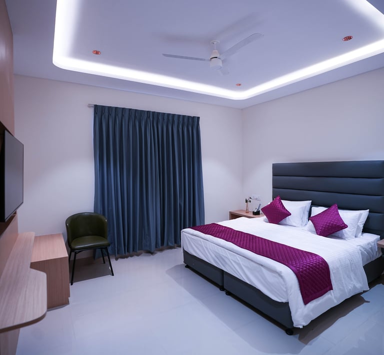 Modern hotel bedroom featuring a king bed with purple accents, cove lighting, and a wall-mounted TV.