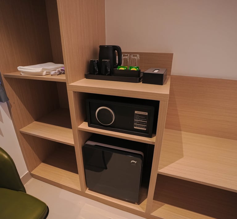 Hotel room amenities featuring a digital safe, mini fridge, and electric kettle on wooden shelves.