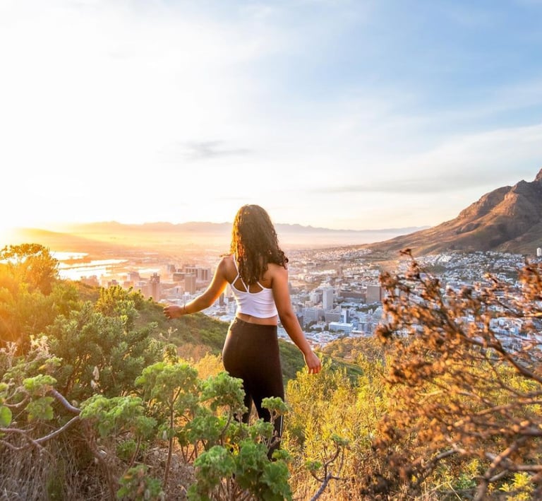 Woman standing between green bushes, looking over Cape Towns CBD. The city is basking in golden morning sunlight. 