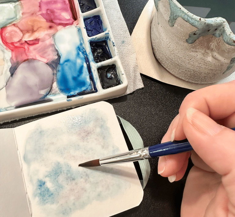 A person holding a paintbrush painting in a skechbook with a watercolor palette in the background.