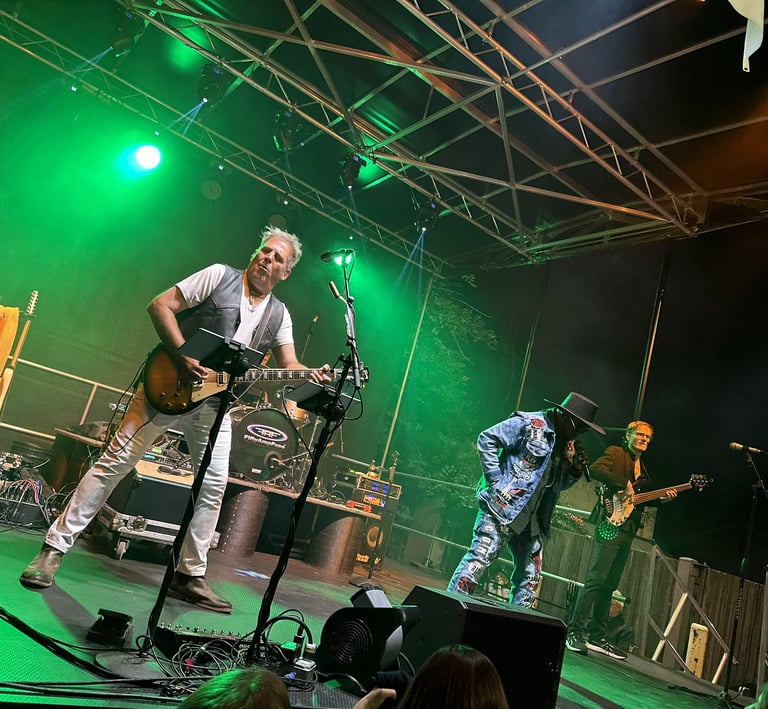 Live rock band performing on an outdoor stage with green lighting and electric guitar player.