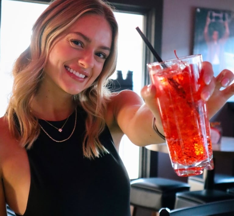 Smiling woman holding a tall iced red cocktail with a cherry and straw at a bar.