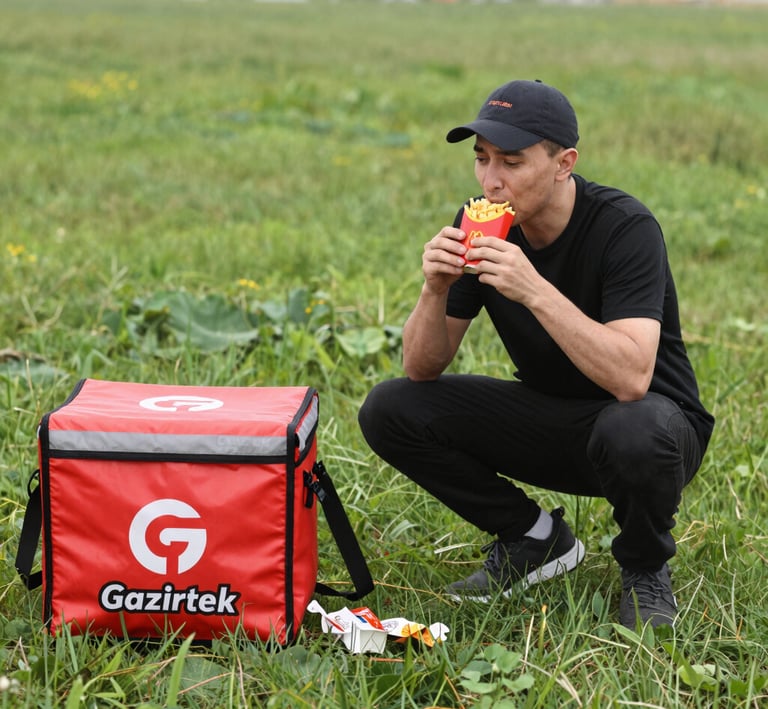 A vibrant delivery driver holding a box filled with fresh ingredients outside a bustling kitchen.