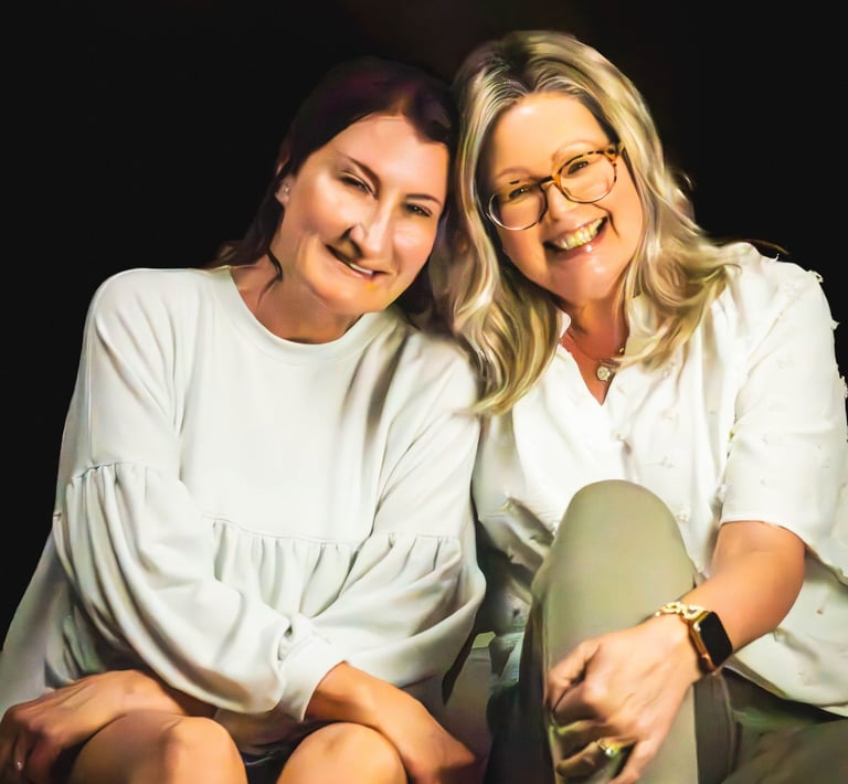 Two smiling women sitting closely together in white shirts against a dark background.