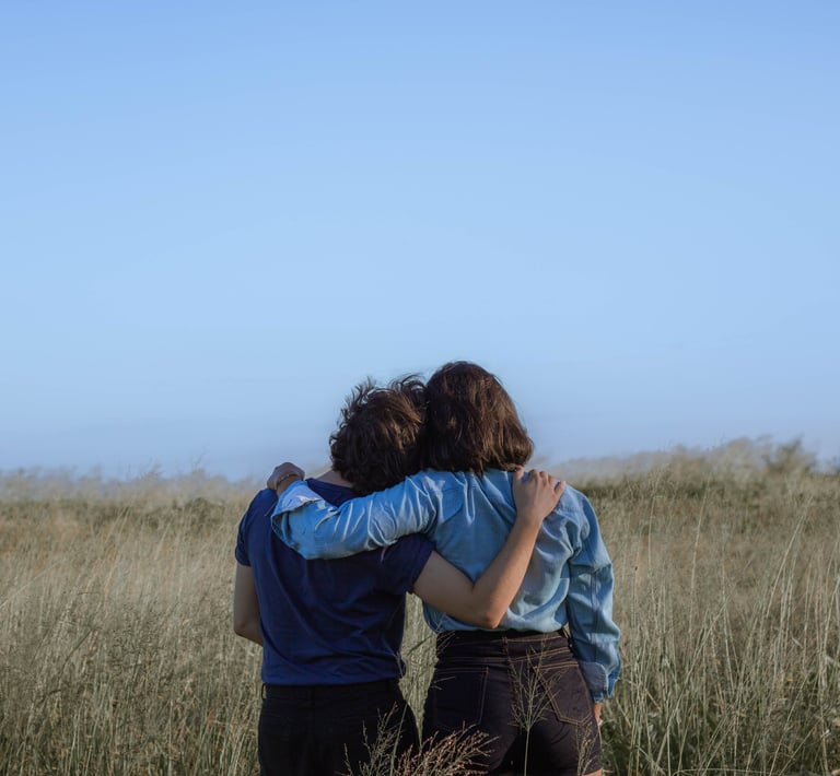 Two friends hugging and looking at the horizon in a grassy field under a clear blue sky.