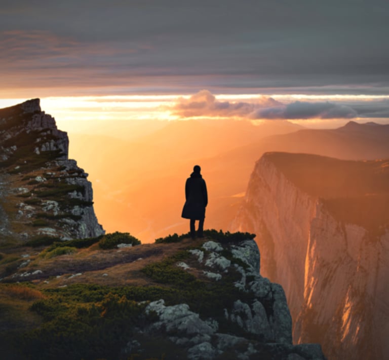A lone person in a dark coat stands on a mountain cliff overlooking a vast, sunlit valley with rugged peaks at sunset