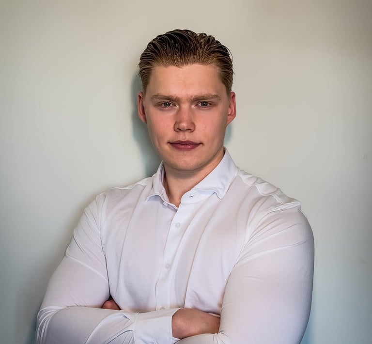 Professional portrait of a young man with crossed arms wearing a white dress shirt against a neutral wall.