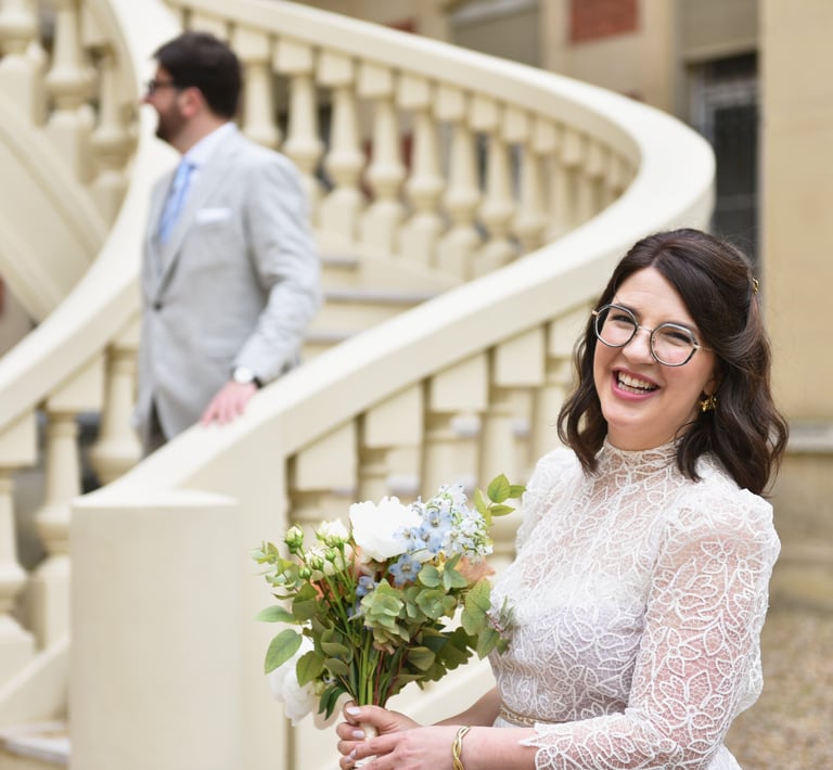 Une mariée sourit face caméra, son mari est derrière sur un escalier il descend