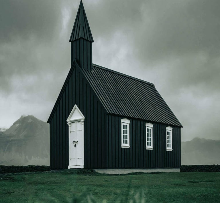 Small black church with white trim and cross-topped steeple set against dramatic cloudy sky.